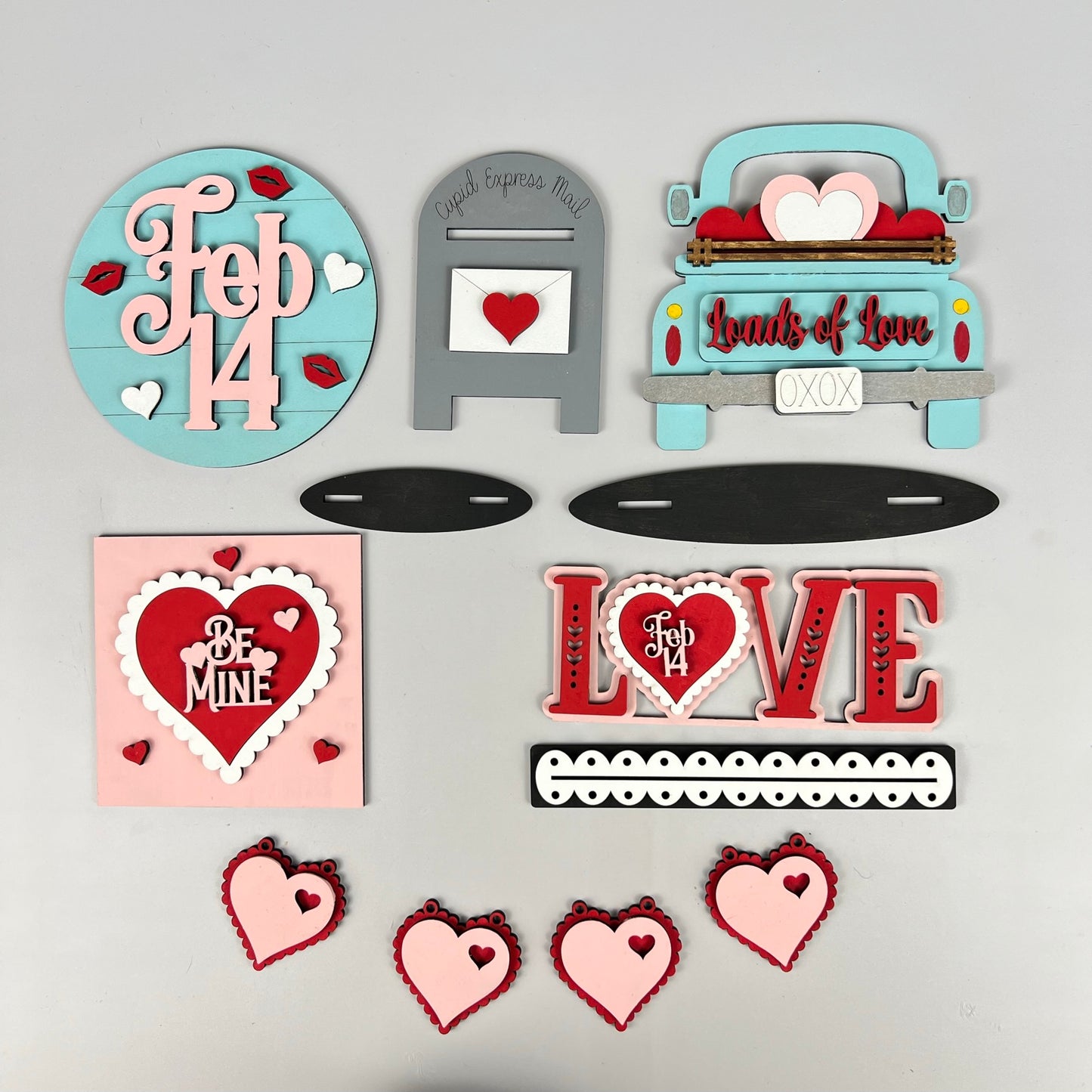 Add a touch of charm to your home décor with Janet's Craft Corner's Loads of Love Tiered Tray Set. This delightful DIY kit includes Valentine's Day-themed wooden decorations such as a blue truck adorned with hearts, a "Cupid Express Mail" mailbox, and lovely phrases like "Loads of Love" and "Be Mine," all featuring red and pink heart designs.