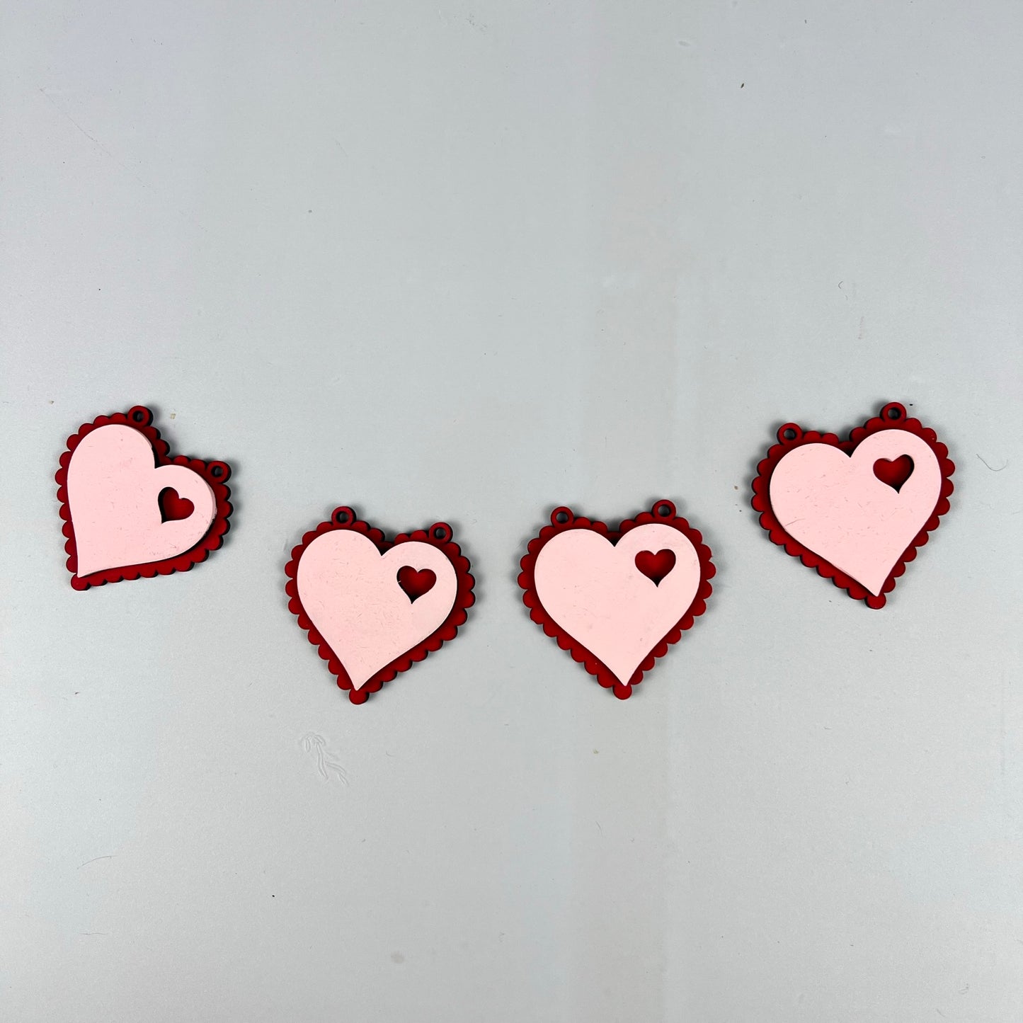 Four pink heart-shaped cutouts, each featuring small red hearts inside and outlined with a scalloped red border, from Janet's Craft Corner's Loads of Love Tiered Tray Set, are arranged in a row on a light gray background, offering the perfect touch of charm to your home décor.