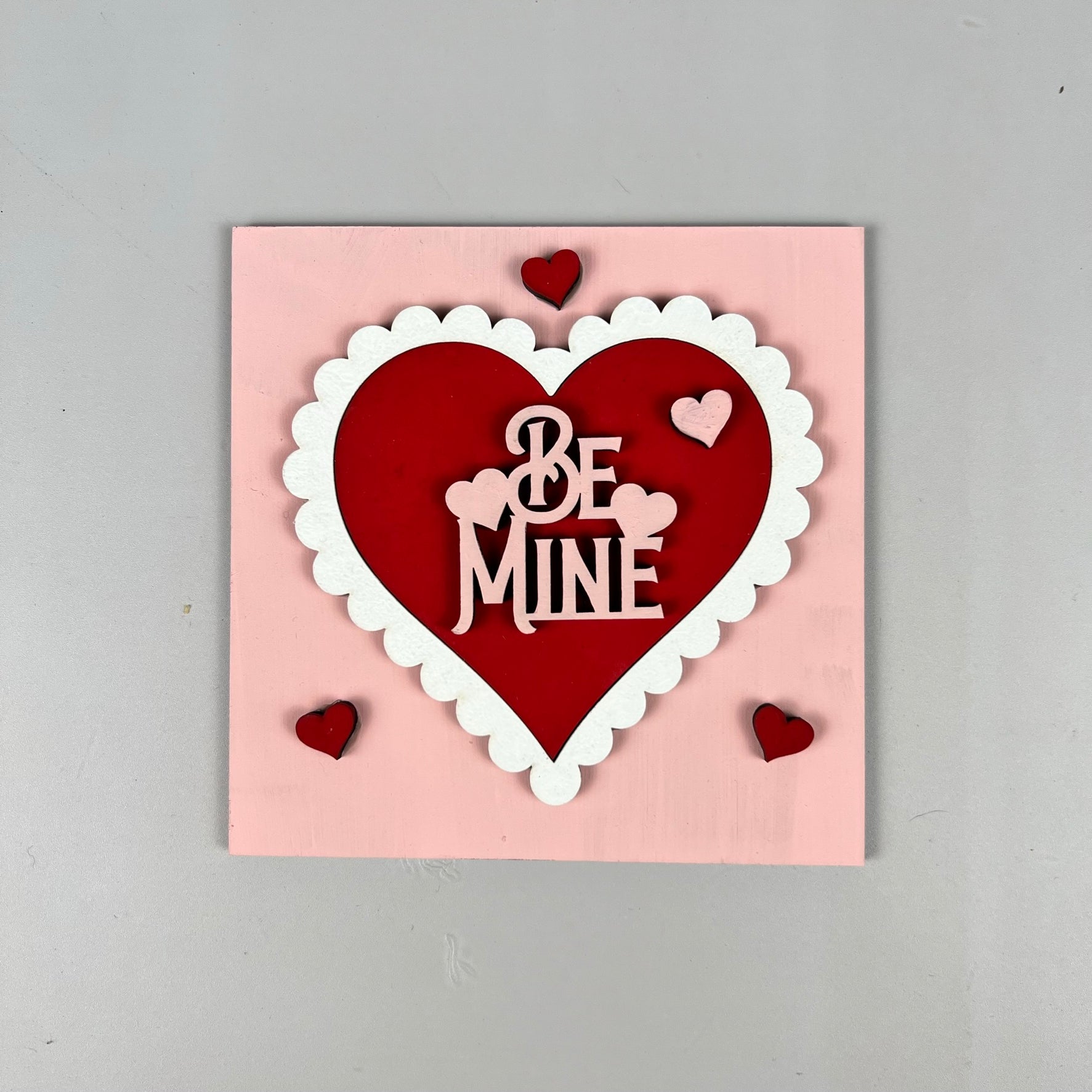 The Loads of Love Tiered Tray Set from Janet's Craft Corner includes a charming pink square card, ideal for DIY kit projects. It showcases a large red heart with a white scalloped border and the phrase "Be Mine" elegantly inscribed in white letters inside the heart. Four smaller red hearts decorate the corners, making it an adorable piece for any home décor ensemble.