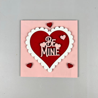 The Loads of Love Tiered Tray Set from Janet's Craft Corner includes a charming pink square card, ideal for DIY kit projects. It showcases a large red heart with a white scalloped border and the phrase "Be Mine" elegantly inscribed in white letters inside the heart. Four smaller red hearts decorate the corners, making it an adorable piece for any home décor ensemble.