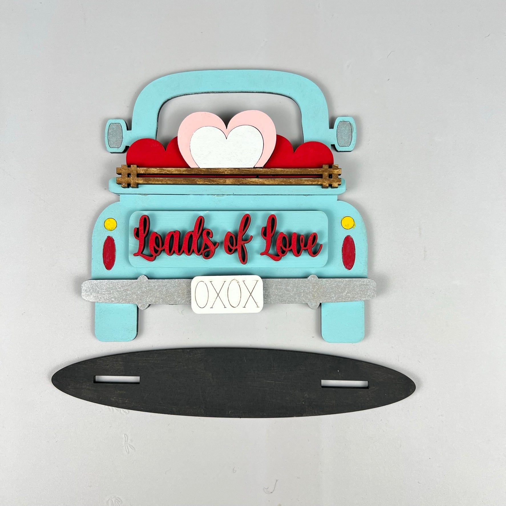 Illustration of a blue truck from the back, loaded with red and white heart cutouts. The phrase "Loads of Love" is featured on the truck. Perfectly designed for inclusion in a tiered tray set, the gray rectangular base below includes two horizontal slots, making it an excellent choice for your home décor or as part of a DIY kit from Janet's Craft Corner's Loads of Love Tiered Tray Set.