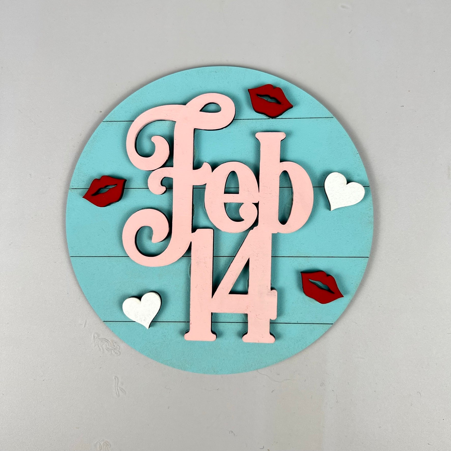 The "Loads of Love Tiered Tray Set" by Janet's Craft Corner features a round sign with "Feb 14" in pink letters on a teal background, accented with red lips and white hearts. It's an ideal addition to your tiered tray arrangement or as a component in a DIY home décor kit for Valentine's Day.