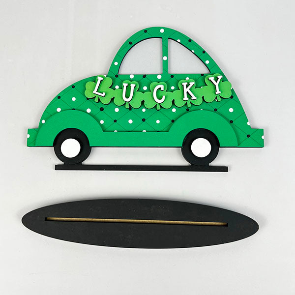 The *Lucky St. Patrick's Volkswagen Shelf Sitter* from Janet's Craft Corner is a decorative green car reminiscent of a Volkswagen, emblazoned with "LUCKY" and adorned with shamrocks, perfect for crafting. It features a separate black oval base for easy attachment, making it an ideal DIY kit for St. Patrick's Day decorations against a simple white background.