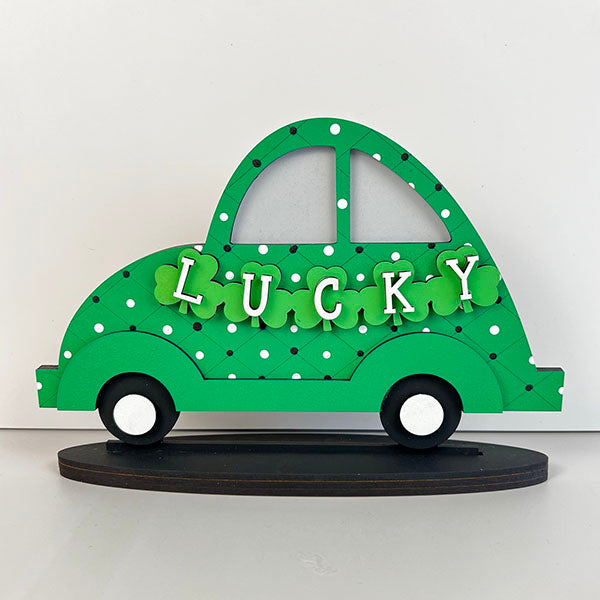 The Lucky St. Patrick's Volkswagen Shelf Sitter by Janet's Craft Corner showcases a decorative green car cutout with a polka dot pattern and the word "LUCKY" across the center, embellished with clover shapes. Ideal for St. Patrick's Day decor, this charming car sits gracefully on a black base.