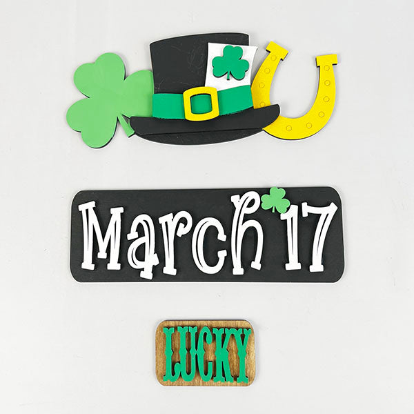 Celebrate St. Patrick's Day with Janet's Craft Corner's March 17 Interchangeable Insert DIY home decor kit, highlighting a black hat decorated with green shamrocks and a yellow horseshoe, a charming sign that reads "March 17," and a versatile wooden block showcasing "Lucky" in bold green letters.