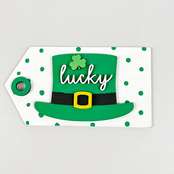 A festive tag from Janet's Craft Corner's Lucky St Patricks Tiered Tray Kit features a green top hat with a shamrock, highlighted by the word "lucky" in white script. The background showcases green polka dots on white, and it includes a small circular hole on the left for hanging with your home décor items.