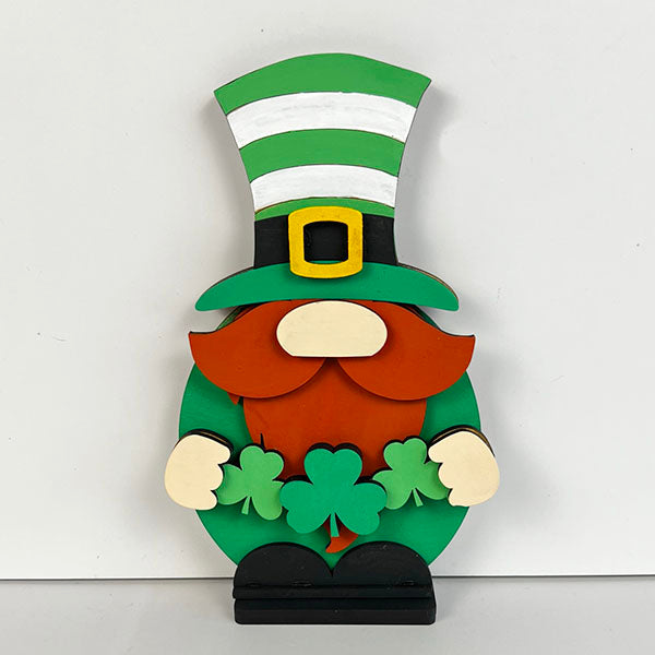 The Leprechan Gnome Shelf Sitter from Janet's Craft Corner features a charming wooden finish, topped with a green and white striped top hat and a bushy red beard. It clutches two green shamrocks, making it a delightful addition to your home décor against a plain white backdrop.