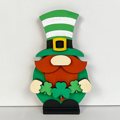 The Leprechan Gnome Shelf Sitter from Janet's Craft Corner features a charming wooden finish, topped with a green and white striped top hat and a bushy red beard. It clutches two green shamrocks, making it a delightful addition to your home décor against a plain white backdrop.