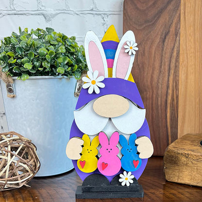 The Easter Peep Gnome by Janet's Craft Corner features a charming design with a purple hat and bunny ears. It's holding three vibrant bunny figures adorned with hearts, making it an ideal enhancement for your Easter decor. Displayed on a table surrounded by a potted plant, wicker ball, and wooden board in the background, this whimsical gnome fits perfectly into any festive scene.