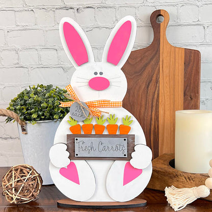 The Carrot Patch Bunny Shelf Sitter from Janet's Craft Corner is an adorable wooden bunny featuring pink ears and a nose, holding a sign adorned with tiny wooden carrots labeled "Fresh Carrots." Ideal for home décor, it is complemented by a plant, candle, and wooden boards set against a brick background.