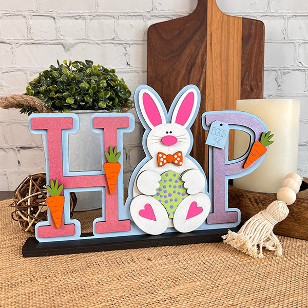 Add a touch of Easter charm to your holiday decor with Janet's Craft Corner's "HOP Standing Bunny Shelf Sitter." This delightful piece features the word "HOP" with a charming bunny in the center, beautifully accented with carrots. The rustic design, complemented by plants and candles in the background, makes it perfect for your festive setup!