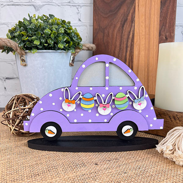 This Easter, the Easter Volkswagen Shelf Sitter from Janet's Craft Corner adds a stylish touch to your home décor. The wooden decoration features a purple car adorned with white polka dots and delightful bunny and egg designs. It is beautifully complemented by a small potted plant, round wicker accents, and a candle in the background.