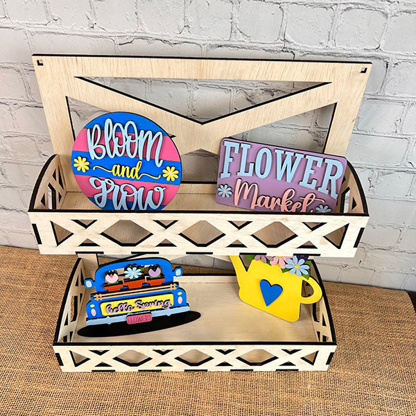 A hand-painted wooden shelving unit with two tiers showcases vibrant decorative items. The top shelf highlights "Bloom and Grow" and "Flower Market" signs, from Janet's Craft Corner's Flower Market Mini Shelf Sitters. The bottom shelf displays a "Happy Spring" sign with a blue truck and a yellow watering can.