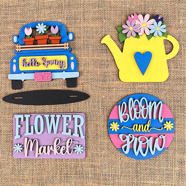 Four vibrant spring-themed wooden signs from Janet's Craft Corner, all hand-painted, are showcased on a burlap background. Included in the set is a blue truck filled with flowers, a yellow watering can decorated with blossoms, the "Flower Market" sign from the Flower Market Mini Shelf Sitters collection, and a round "Bloom and Grow" sign featuring floral accents.