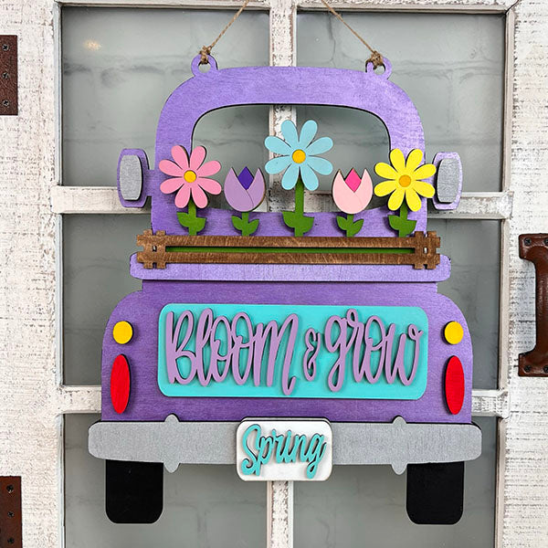 Bring charm to your home décor with Janet's Craft Corner's Spring Blooms Interchangeable Insert. This delightful DIY home decor kit features a hand-painted purple truck brimming with vibrant flowers and a whimsical "Bloom & grow" message. A quaint little "Spring" sign dangles from the bumper, all beautifully set against a rustic white door backdrop.