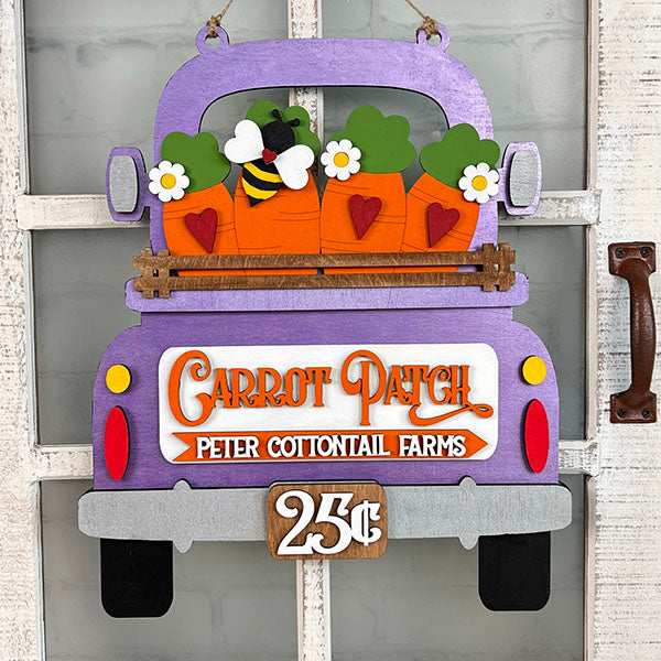 Enhance your home decor or DIY craft kit with the Carrot Patch Interchangeable Insert by Janet's Craft Corner. This decorative purple truck sign, featuring flowers, bees, and carrots along with the text "Carrot Patch Peter Cottontail Farms" and "25¢," is perfect for hanging against a rustic white wooden background.