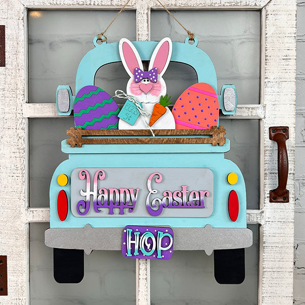 This charming Easter Bunny Interchangeable Insert from Janet's Craft Corner, an ideal DIY home decor kit, showcases a lively blue truck carrying a bunny, eggs, and a carrot. With "Happy Easter" and "Hop" embellishments, it beautifully adorns a rustic white door.