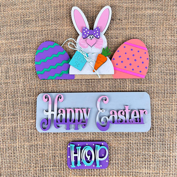 This interchangeable home decor kit from Janet's Craft Corner features a decorative Easter sign on burlap, displaying a pink-eared bunny clutching a carrot amidst colorful eggs. With text panels that say "Happy Easter" and "Hop," it adds vibrant charm to your space, making it perfect as an Easter Bunny insert for your seasonal displays.