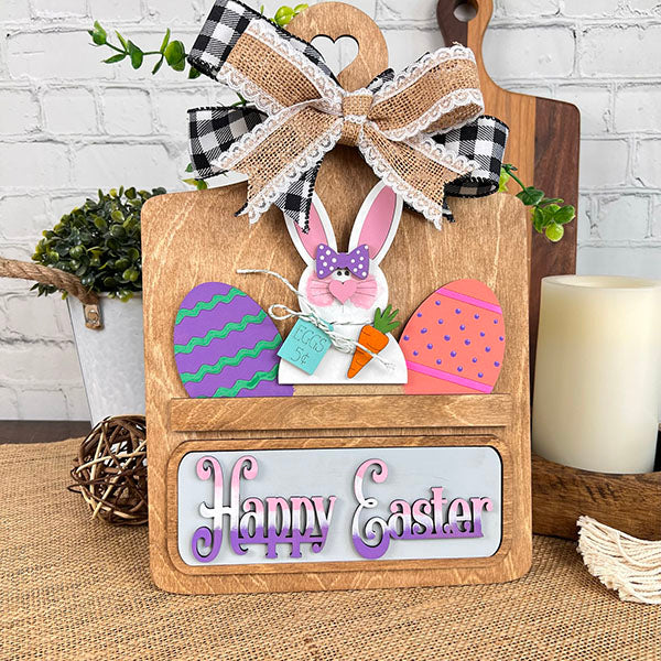 Janet's Craft Corner presents the Easter Bunny Interchangeable Insert, a delightful DIY home decor kit. This wooden decoration showcases a bunny with glasses and a bow tie holding a carrot, accented by colorful eggs and topped with a checkered bow. The "Happy Easter" sign below enhances its charm against a rustic background.