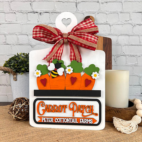 Decorative sign from Janet's Craft Corner, featuring a checkered red ribbon and three carrots adorned with bees, flowers, and hearts. Includes the text "Carrot Patch" and "Peter Cottontail Farms," set against a brick wall. Ideal for DIY home decor enthusiasts using the Carrot Patch Interchangeable Insert kit. Complete your personalized look with candles and plants.