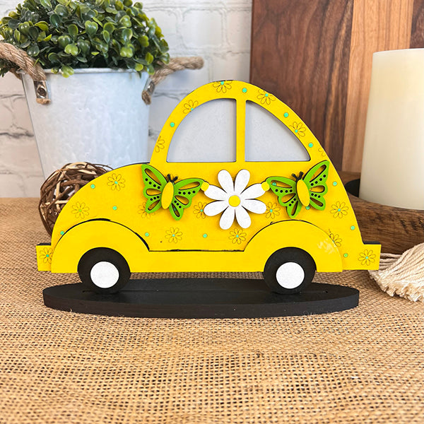 The "Spring Volkswagen Shelf Sitter" by Janet's Craft Corner features a decorative wooden cutout of a yellow Volkswagen, accented with two green butterflies and a white flower, all perched on a dark oval base. Ideal for enhancing home décor, it is displayed on a wooden table against a white brick background with a plant in the corner.