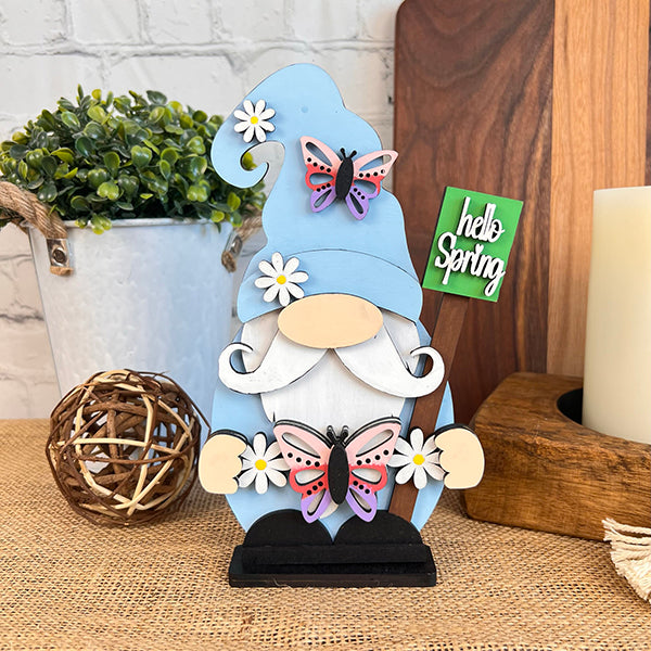 A decorative Spring Butterfly Gnome from Janet's Craft Corner, featuring a blue hat and holding two butterflies amidst daisies, is displayed on a table. Beside it lies a charming "Hello Spring" sign, complemented by a potted plant, wicker ball, and candle in the background—ideal for enhancing your home décor.