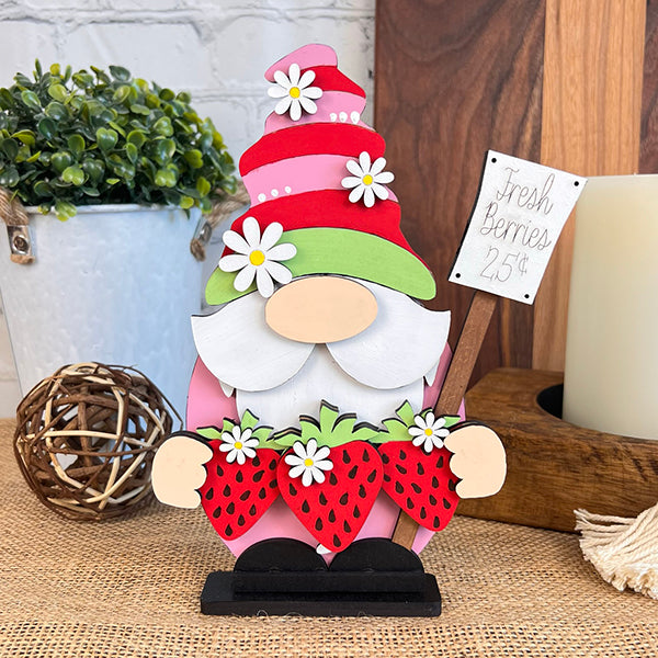 The delightful Strawberry Gnome from Janet's Craft Corner features a red hat with green trim and is decorated with white flowers while holding large red strawberries. Ideal for farmhouse décor, it stands on a burlap surface next to a potted plant, woven ball, and candle, accompanied by a sign that reads "Fresh Berries 25¢.