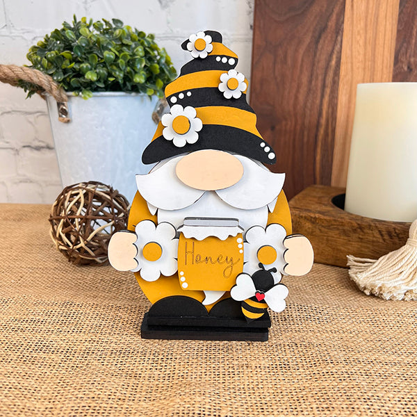 The Bee Gnome from Janet's Craft Corner, a delightful piece for farmhouse decor, showcases a floral-adorned yellow and black hat. It holds a "Honey" sign and stands on woven fabric with a small bee and decorative items in the background.