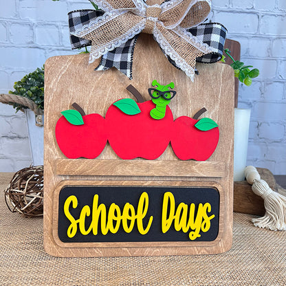 Featuring three red apples and a cartoon green worm with glasses above the words "School Days" in yellow, the School Days Interchangeable Insert by Janet's Craft Corner is perfect for farmhouse decor. This DIY home decor kit is topped with a gingham and burlap bow, beautifully set against a brick wall background.