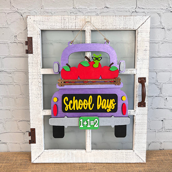 Introducing the "School Days Interchangeable Insert" DIY home decor kit by Janet's Craft Corner. This beautifully designed craft kit features a decorative sign with a purple truck set against a white rustic frame, ideal for farmhouse decor. The truck is whimsically adorned with red apples and displays the phrases "School Days" and "1+1=2" in vibrant yellow and green, all against a light gray brick wall backdrop.