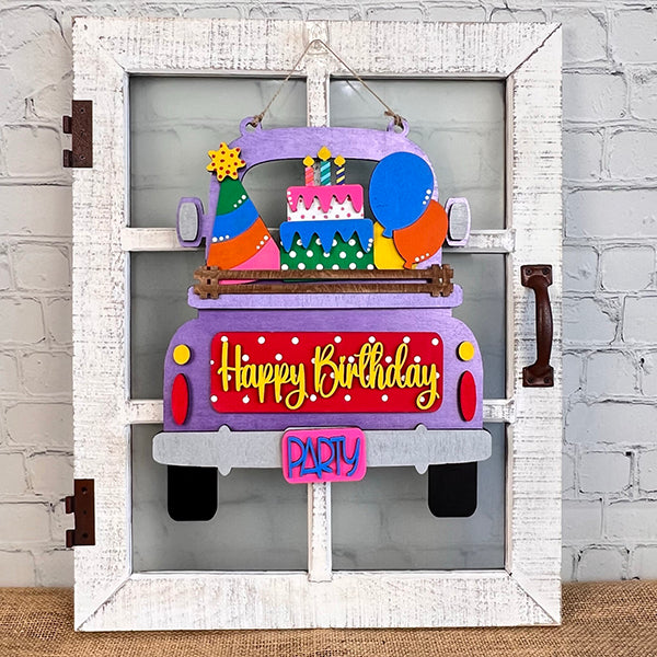 The "Birthday Interchangeable Insert" from Janet's Craft Corner features a vibrant wooden design of a truck with "Happy Birthday" on its tailgate, carrying a cake, balloons, and party hats. This DIY home decor kit is designed to hang on a white window frame against a brick wall for a festive touch.