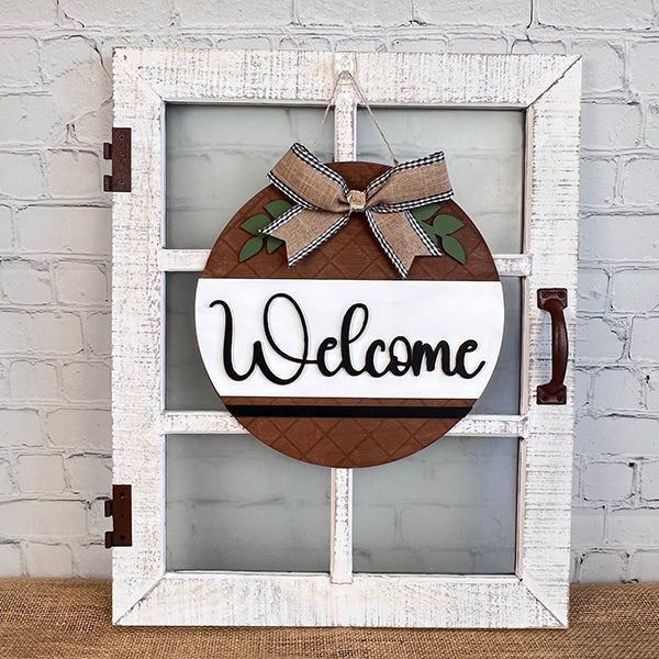 A decorative piece from Janet's Craft Corner, this Welcome Round Door Hanger with a circular design reading "Welcome" is perfect for farmhouse decor. It hangs on a rustic white vintage window frame that features an antique handle. The hanger showcases a brown quilted pattern, adorned with a checkered ribbon bow and small green leaves.