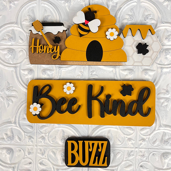 Introducing the Bee Interchangeable Insert - Painted from Janet's Craft Corner. This bee-themed wall decoration features a captivating beehive with a bee and honey pot, the phrase "Bee Kind" surrounded by delicate flowers, and the word "Buzz" against a stylish white patterned background. Customize your decor effortlessly with interchangeable craft inserts for any occasion.