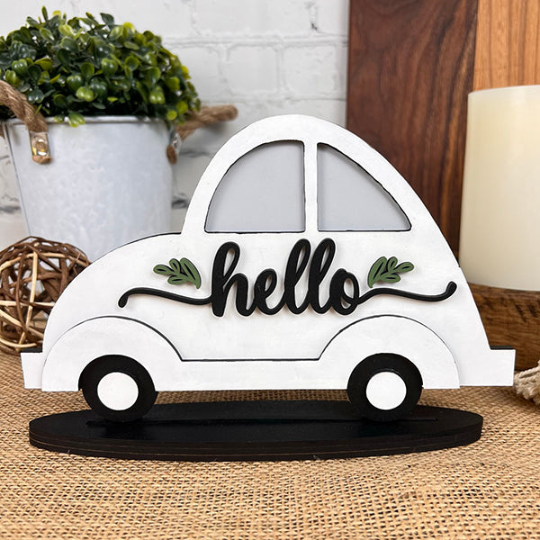 The "Hello Volkswagen" from Janet's Craft Corner is a decorative wooden cutout in the shape of a car, ideal for farmhouse decor. It features the word "hello" in elegant black script on its side. This delightful shelf sitter is displayed on a table with a small plant, wicker ball, and candle adorning the background.