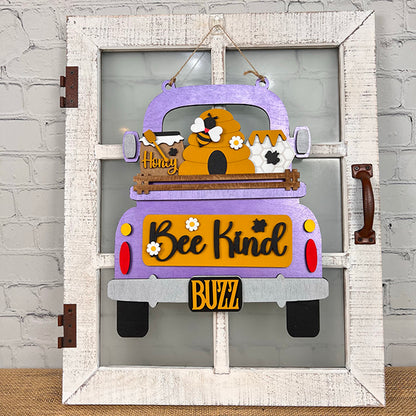 A decorative sign featuring the Bee Interchangeable Insert from Janet's Craft Corner presents a hand-painted purple truck with a beehive and bees. The piece, which prominently displays "Bee Kind" and a "BUZZ" license plate, stands out against a textured gray wall and is enhanced by the brown handle on its white wooden frame.