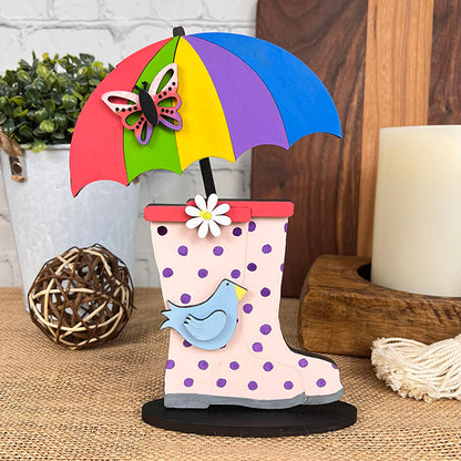 Introducing a vibrant decorative arrangement showcasing polka dot rain boots, adorned with a blue bird and topped by a rainbow umbrella, butterfly, and flower. This charming scene is complete with the Spring Standing Signs from Janet's Craft Corner, enhanced by a candle, woven sphere, and potted plant on a wooden surface to elevate your home décor.