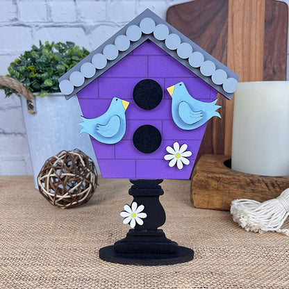 The "Spring Standing Signs" from Janet's Craft Corner is a decorative purple birdhouse ideal for home décor. It showcases two blue birds and white flowers upon a black pedestal, enhanced by the addition of a plant, wooden decor, and candle on a burlap surface to evoke a delightful Spring atmosphere.