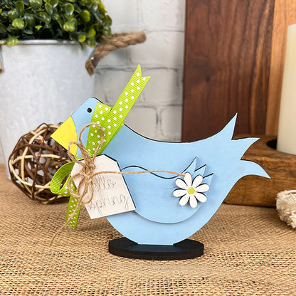 A charming piece from Janet's Craft Corner, the Spring Standing Signs include a blue wooden bird figurine with a yellow beak and green ribbon, prominently displayed on a burlap surface. Featuring delightful flower details and a tag reading "Hello Spring," it adds an inviting touch to your home décor. Surrounding DIY kit elements like potted plants and decorative items complete the scene with grace.