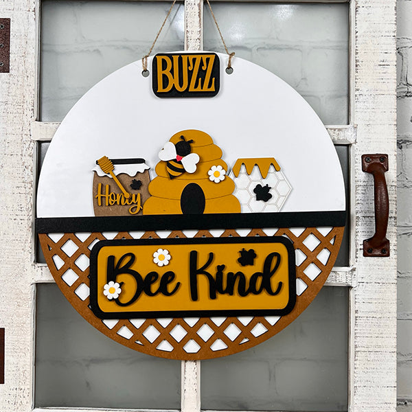 Discover the Bee Interchangeable Insert - Painted from Janet's Craft Corner. This round wooden decorative hanging sign features a charming beehive, bees, and honeycomb motif. It includes the words "Buzz," "Honey," and "Bee Kind" with a lattice pattern at the bottom, offering versatile decor options with its interchangeable bee inserts.