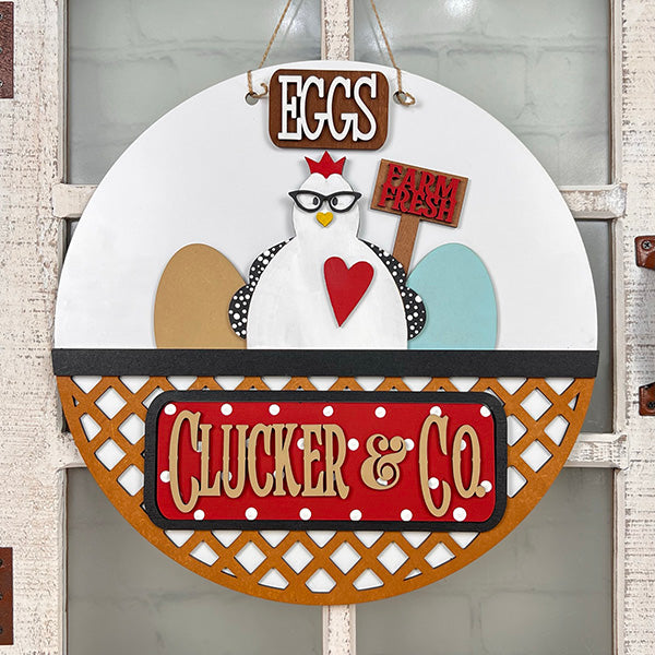 Janet's Craft Corner introduces the Clucker & Co. Interchangeable Insert - Painted, a decorative piece featuring a delightful chicken motif adorned with glasses, alongside a red heart and colorful eggs. The sign displays "EGGS" and "Farm Fresh" at the top, with "Clucker & Co." beneath on a rustic lattice background, making it an ideal choice for farmhouse decor enthusiasts seeking versatile craft inserts.