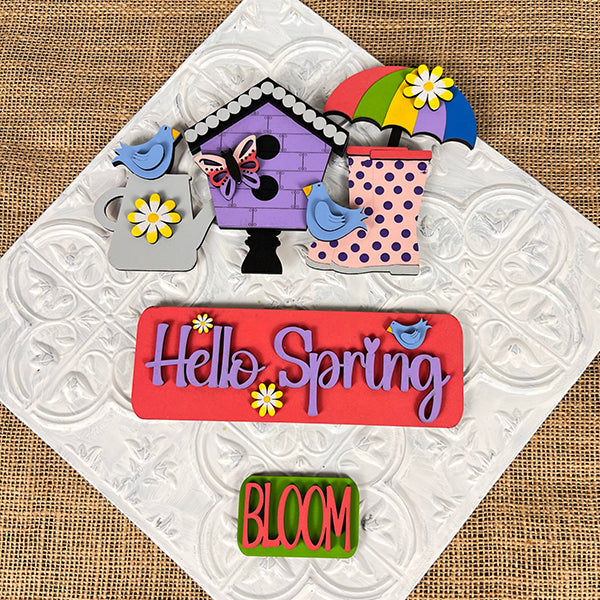 Complete your textured surface with vibrant spring-themed decorations from Janet's Craft Corner. The scene includes a birdhouse, bluebirds, an umbrella, and rain boots. A charming sign displays "Hello Spring" in purple adorned with flowers. This delightful setup is part of the Hello Spring Interchangeable Insert DIY home decor kit, featuring an additional option that says "BLOOM.