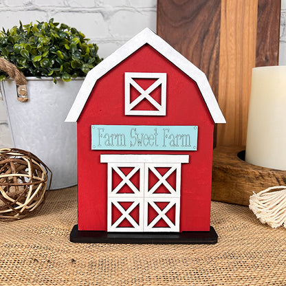 The "Farm Standing Shelf Sitter" from Janet's Craft Corner introduces a farmhouse decor vibe with its decorative red barn and white trim, complete with a sign saying "Farm Sweet Farm." Set on a burlap surface, it enhances the rustic feel with accompanying decor items like a potted plant, woven ball, and candle, crafting an inviting DIY kit atmosphere.