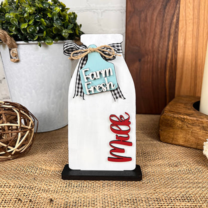 The Farm Standing Shelf Sitter by Janet's Craft Corner embodies farmhouse charm with its milk bottle shape, decorated with a "Farm Fresh" blue tag and "Milk" in bold red letters. The piece is completed with a black and white bow at its black base and is perfectly complemented by rustic decor such as a wooden board, a plant, and a woven ball.