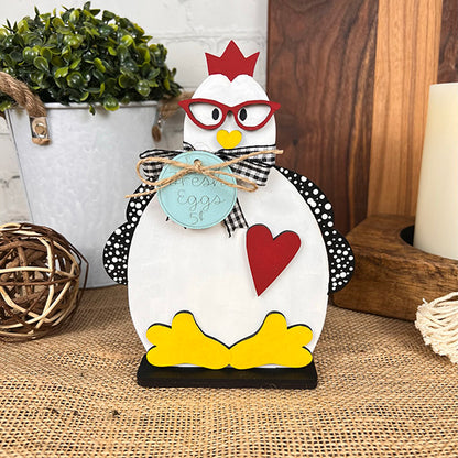 The Farm Standing Shelf Sitter from Janet's Craft Corner is a quaint wooden chicken figure featuring a red crown, glasses, and a heart on its side. It stands with yellow feet and is adorned with a checkered bow while holding a "Fresh Eggs" sign. This piece is ideal for enhancing your farmhouse decor with its touch of rustic charm amidst plant and vintage-inspired elements.