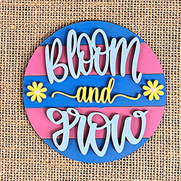 A hand-painted round sign with "Bloom and Grow" in cursive beautifully adorns a burlap surface, featuring a background of alternating pink and blue stripes. This design, highlighted by two bright yellow flowers, evokes the charm of the Flower Market Mini Shelf Sitters by Janet's Craft Corner.