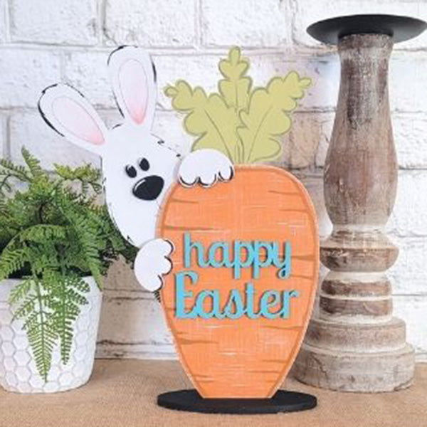 The "Bunny and Carrot Happy Easter Shelf Sitter" from Janet's Craft Corner features a charming Easter bunny peeking out from behind a large DIY carrot inscribed with "happy Easter." This piece, part of the Bunny and Carrot collection, is beautifully displayed on a surface decorated with a lush plant in a white pot and complemented by a rustic candle holder nearby.