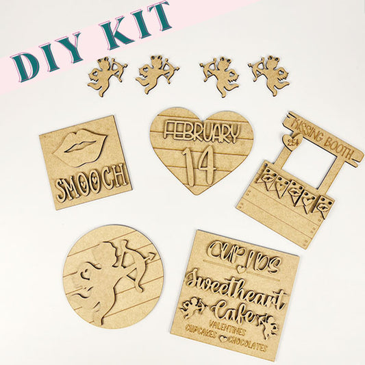 The Cupid Kissing Booth Tiered Tray Kit from Janet's Craft Corner provides wooden Valentine's Day decor pieces, including cupid shapes and a heart featuring "February 14." Create your own Cupid Kissing Booth and "Cupid's Sweetheart Cafe" with a square marked "Smooch" alongside other delightful designs.
