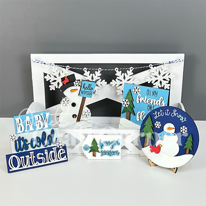 A festive winter display featuring a variety of snowman-themed decorations, snowflake garlands, and wintery signs like "Baby, it's cold outside." The "Hello Winter Tiered Tray Kit" from Janet's Craft Corner adds a charming touch to the home decor with its blue, white, and green elements.