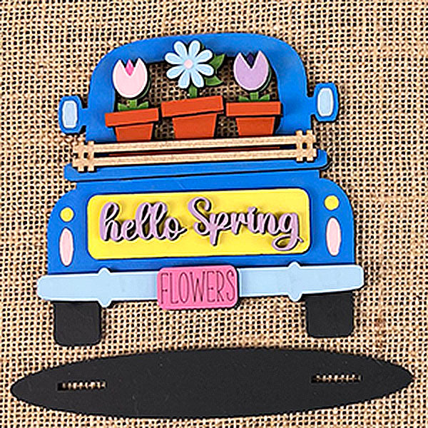 A decorative sign, inspired by Janet's Craft Corner's Flower Market Mini Shelf Sitters, features a blue truck adorned with flower pots overflowing with tulips and daisies. The "FLOWERS" license plate enhances the "hello Spring" message on the tailgate set against a rustic burlap backdrop.