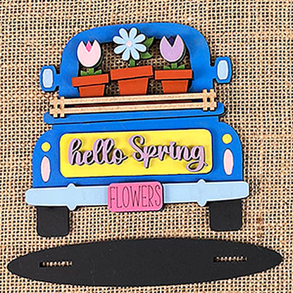 A decorative sign, inspired by Janet's Craft Corner's Flower Market Mini Shelf Sitters, features a blue truck adorned with flower pots overflowing with tulips and daisies. The "FLOWERS" license plate enhances the "hello Spring" message on the tailgate set against a rustic burlap backdrop.